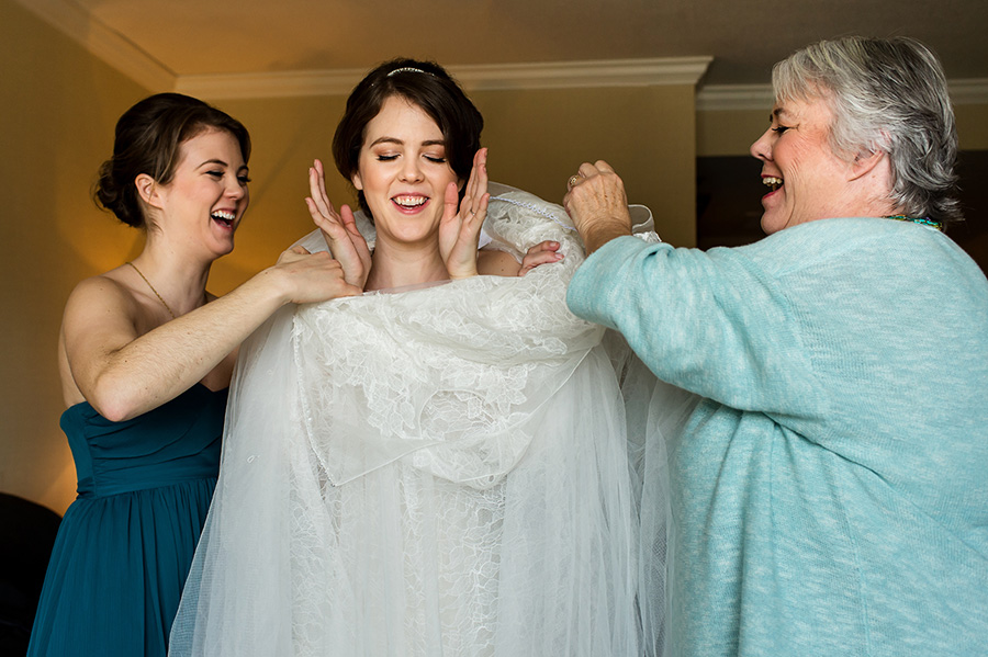 Maid of honor and mother of the bride helping bride into her wedding gown.