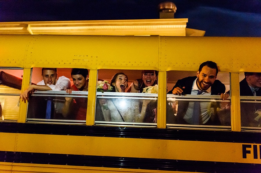Bride, groom and guests hanging out of school bus windows.