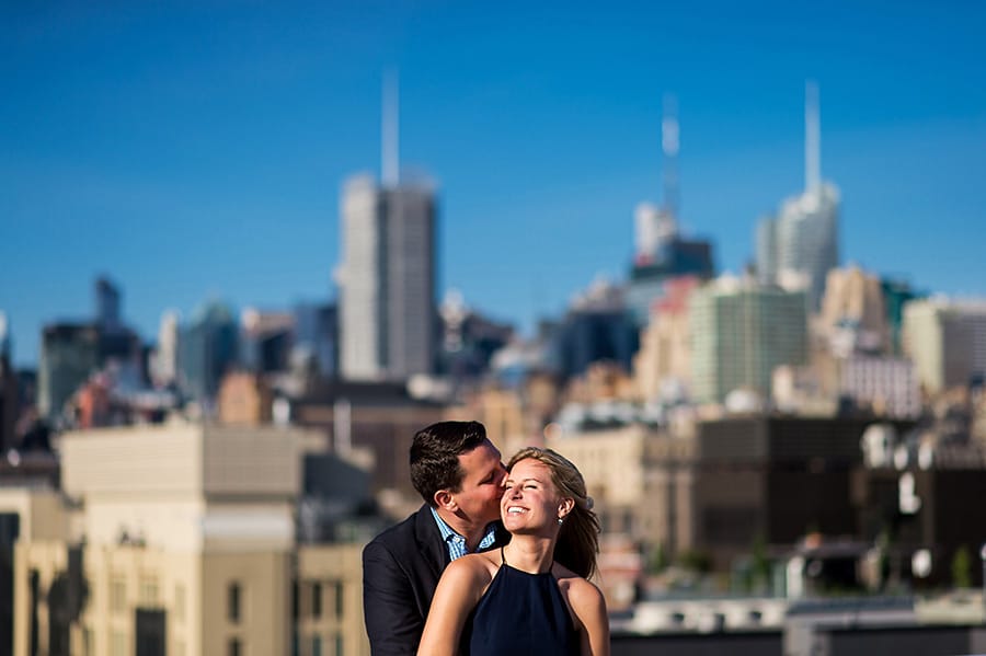 Engaged bride-to-be smiling as groom to be kisses her on the cheek with NYC skyline in background.